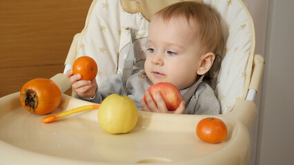 Fototapeta na wymiar Portrait of cute baby boy sitting in highchair and holding fresh tasty fruits