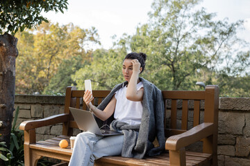 Curly and pretty freelancer in warm jacket having video call on smartphone near laptop, coffee to go and orange on wooden bench in park at daytime in Barcelona, Spain