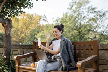 Cheerful young freelancer in warm jacket and earphones waving hand at smartphone while having video call on smartphone near laptop, coffee to go and orange on wooden bench in park in Barcelona, Spain