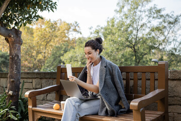 Fototapeta premium Smiling young freelancer in warm jacket and earphones smiling, covering mouth, having video call on smartphone near laptop and coffee to go on wooden bench in park in Barcelona, Spain