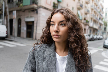 Brunette and pretty young woman in casual jacket looking away while standing on blurred city street...