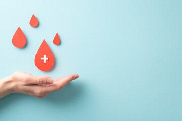 First person top view of a female hand with blood drops falling onto her palm on a pastel blue background for World Blood Donor Day
