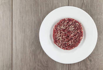 Raw hamburger steak with sesame seeds on white plate over wooden background.