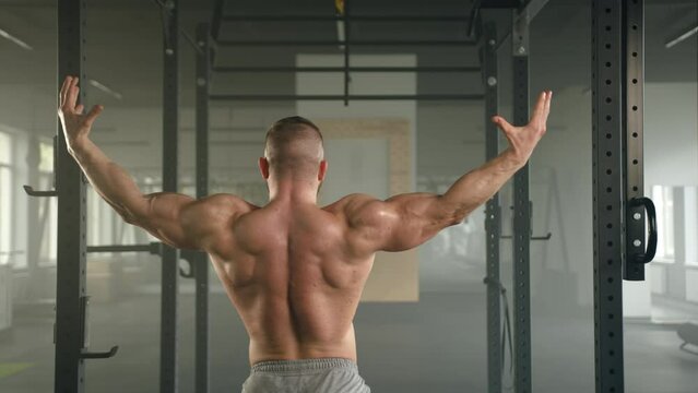 Medium shot back view of bodybuilder showing his muscular back, biceps and triceps. Man with naked torso flexes arms with fists. Power rack in focus. High quality 4k footage