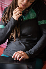 Woman in her forties talking on the phone and typing on the computer on the sofa at home, woman with dreadlocks, glasses, piercings. Large monstera in the background.