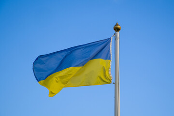 One Ukrainian blue and yellow flag is hanging on a flagpole against a clear sky. Ukrainian flag waving in the wind against the blue sky.
