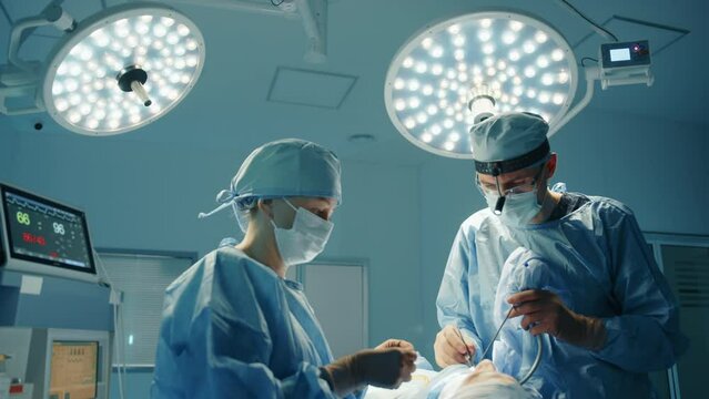 Young male surgeon and nurse performing plastic surgery procedure on face. Spacious operating room, led operation theatre lights, monitor in background. High quality 4k footage
