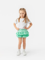 A 5-year-old girl with long hair stands with her hands folded on her belt in a white T-shirt, green skirt on a white background.