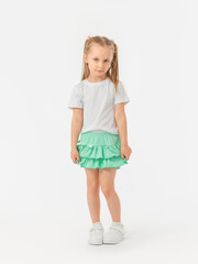 A beautiful serious 5-year-old girl with long hair is standing in a T-shirt and a skirt-shorts, looking at the camera with her leg bent at the knee in sneakers on a white background.