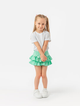 A Caucasian girl with long hair 5 years old in a T-shirt and a green skirt stands with her arms and legs crossed in sneakers on a white background. Looking into the camera