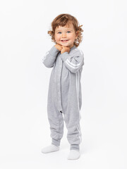 A happy 2-year-old toddler with curly hair in a gray jumpsuit and socks stands and smiles with his hands folded under his chin on a white background.