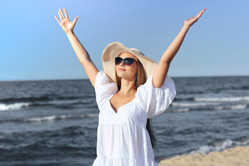 Fototapeta na wymiar Happy blonde woman is on the ocean beach in a white dress, sunglasses and hat, raising hands.