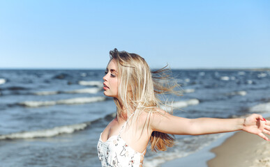 Fototapeta na wymiar Happy blonde beautiful woman on the ocean beach standing in a white summer dress, raising hands.