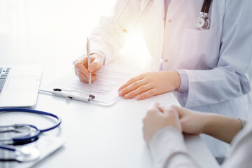 Doctor and patient sitting at the table in clinic. The focus is on female physician's hands filling...