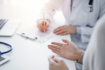 Doctor and patient sitting at the table in clinic. The focus is on female physician's hands filling up the medication history record form or checklist, close up. Medicine concept.