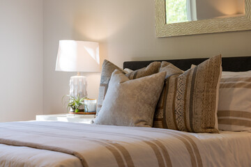 Relaxing bedroom detail of large bed with tan and white bedding, decorative lamp, and soft window light.