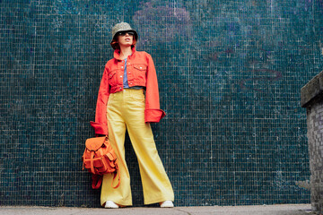 Hipster young woman in bright clothes, sun glasses, backpack bag and bucket hat posing on green...