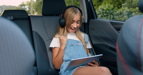 Little girl with headphones playing game and celebrates the victory with digital tablet while sitting in the back seat of car.