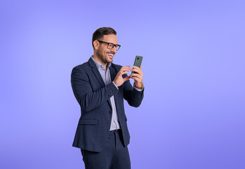 Smiling handsome young entrepreneur wearing eyeglasses and suit text messaging online over smart phone. Happy elegant male CEO telecommuting with cellphone while standing on blue background