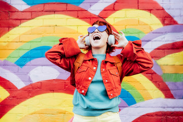 Emotional stylish woman in bright clothes wearing wireless headphones, listening to music and singing on rainbow graffity on the brick wall background. Fashionable hipster lifestyle. - obrazy, fototapety, plakaty
