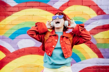 Foto op Canvas Emotional stylish woman in bright clothes wearing wireless headphones, listening to music and singing on rainbow graffity on the brick wall background. Fashionable hipster lifestyle. © okrasiuk