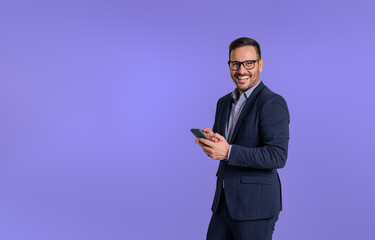 Portrait of elegant male professional scrolling social media happily over mobile phone and looking at camera. Young handsome manager smiling and using cellphone isolated against blue background