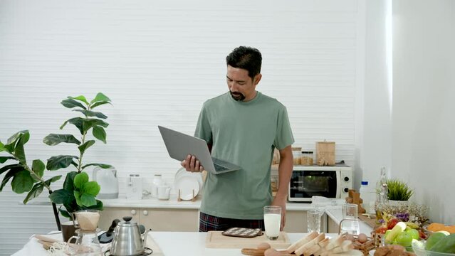 Asian man wearing casual clothes as work clothes at home holds laptop computer hand walk into kitchen find breakfast, Grab cookie put it in mouth, looked down at laptop computer walked out kitchen.