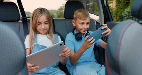 Beautiful children boy and girl having fun playing games on phone and tablet while sitting in a modern car. Little brother and sister are laughing and happily celebrating vin in online game by giving