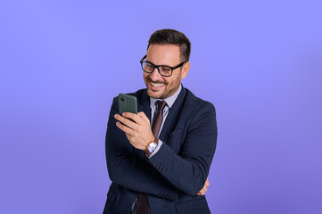 Smiling handsome business manager text messaging over smart phone while standing against blue background. Portrait of cheerful male professional checking social media applications over cellphone