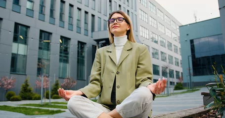 Schilderijen op glas Young caucasian woman in glasses relaxing, sitting in the lotus position on the bench of the courtyard of office building or university. Healthy lifestyle, outdoors © serg