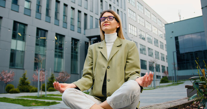 Young caucasian woman in glasses relaxing, sitting in the lotus position on the bench of the courtyard of office building or university. Healthy lifestyle, outdoors