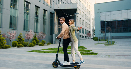 Young cheerful caucasian couple having fun riding electric scooter in sunny weather in the city park near modern office building. Lifestyle. Outside © serg