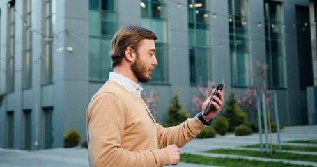 Side view of bearded smiling handsome man talking at mobile phone walking in the street to the office building. Outdoors