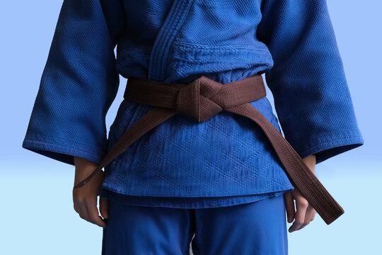 Judo Girl With Brown Belt. A Judoka Teenager Fighter Poses In A Blue Kimono On A Plain Background. Japanese Martial Art. The Process Of Martial Arts Training. Healthy Lifestyle, Sports Concept
