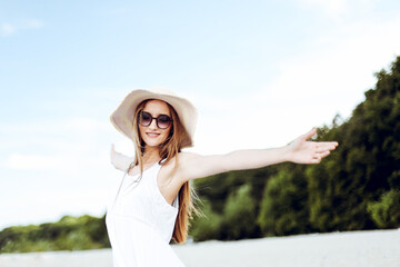 Happy smiling woman in free happiness bliss on ocean beach standing with a hat, sunglasses, and open hands. Portrait of a multicultural female model in white summer dress enjoying nature during travel