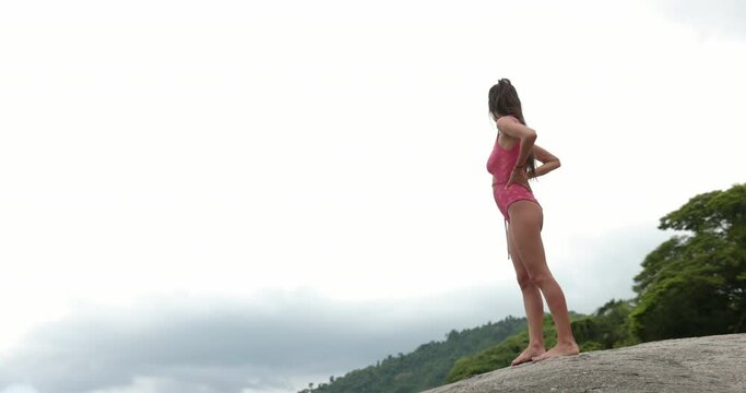 Woman stands on rocks in bikini overlooking tropical destination on overcast day - wide shot