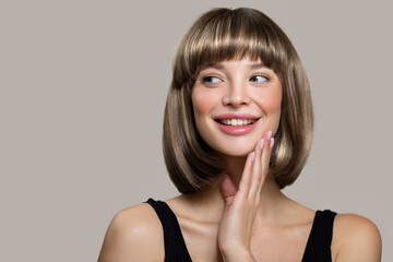 Happy young woman with bob hairstyle smiling while touching her face with her hand. Gray background - obrazy, fototapety, plakaty