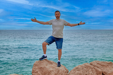 A man stands on a stone against the background of the sea with his arms outstretched