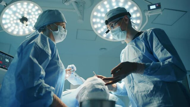 Male young surgeon gives instructions to nurse during plastic surgery operation. Patient is under anaesthetic. Led operation theatre light in focus. High quality 4k footage