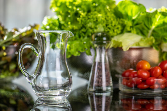 Small clean transparent glass jug on glass table with salad dish green vegetable black ground in kitchen.