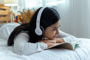 Young woman in pleasure white casual cloth lying down reading book and listen music by headphone on bed. Asian girl imagine story and smiling about novel book.