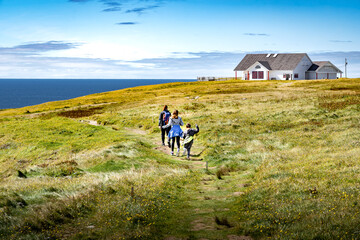Family on vacation running across a grassy field overlooking high cliffs and the Atlantic Ocean at Cape St. Mary's Ecological Reserve Newfoundland Canada. - obrazy, fototapety, plakaty