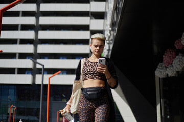 Fototapeta na wymiar Young diverse female with short dyed hair walking outdoor and using a mobile app on a smartphone. Stylish white woman in leopard print clothes walks in the city center with a glass bottle of water