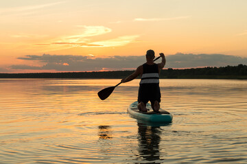 A man in shorts with his back to the camera on his knees on a SUP board with an oar swims in the lake against the background of the sunset sky.