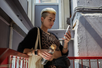 Diverse tattooed woman with short hair using a smart phone with a glass bottle of water in hand and tote bag on shoulder. Sustainability and diversity concept