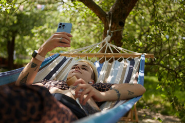 Young diverse woman with short hair lying on a hammock in a park and browsing mobile app on a smartphone. Stylish tattooed female relaxing in a green garden on weekend in summer - obrazy, fototapety, plakaty