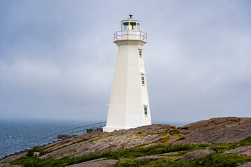 Tall white lighthouse on a rocky shoreline overlooking the Atlantic Ocean during a foggy day along the East Coast Trail at Cape Spear Newfoundland Canada.