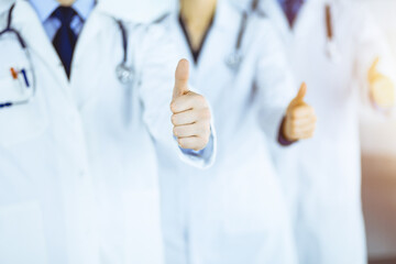 Group of unknown doctors stand as a team with thumbs up in a sunny hospital office. Physicians ready to examine and help patients. Medical help, insurance in health care, best desease treatment and me