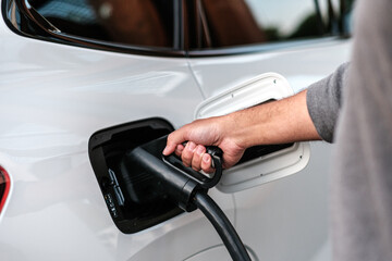 Person plugging in a charging cable to a white car at a electric vehicle fast charging station,...