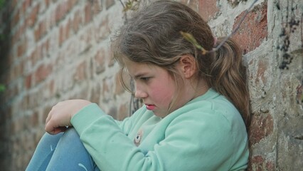 Obraz na płótnie Canvas Upset Frustrated Depressed Young Caucasian Girl Sitting Alone Outdoors Against Brick Wall Hiding from People Parents and Friends